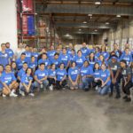 A large group of people wearing matching blue T-shirts pose for a group photo in the Los Angeles Regional Food Bank's warehouse. Many are smiling, standing close on the concrete floor, surrounded by tall shelves and industrial equipment in the background.