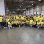 A large group of people in yellow T-shirts poses together in a spacious, warehouse-like setting at the Los Angeles Regional Food Bank. They seem gathered for a community event or activity, standing on a polished concrete floor.
