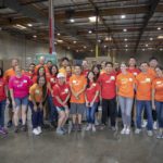 A group of people wearing red and orange t-shirts poses for a photo in a large warehouse, with stacks of boxes behind them. They appear to be volunteering at the Los Angeles Regional Food Bank or participating in an event.