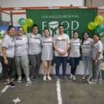 A group of people wearing matching "Deloitte" shirts stand together, smiling in front of a balloon-decorated Los Angeles Regional Food Bank backdrop. They appear to be participating in a volunteer event.