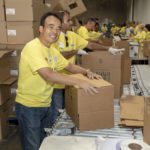Volunteers in yellow shirts diligently pack and organize boxes at the Los Angeles Regional Food Bank, placing items into cardboard boxes on a conveyor belt within the bustling warehouse setting.