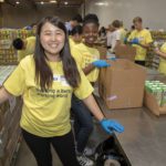A group of volunteers in yellow T-shirts and blue gloves pack boxes with food items at the Los Angeles Regional Food Bank warehouse. A woman in the foreground smiles at the camera, while others work diligently in the background.