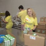 Volunteers in yellow shirts pack food items at the Los Angeles Regional Food Bank warehouse. A woman in the foreground, smiling, holds a carton. Others fill boxes around her, working diligently at a table surrounded by packed boxes and cartons.
