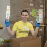 A volunteer in a yellow T-shirt and blue gloves holds two boxes of food at the Los Angeles Regional Food Bank, standing amid large cardboard boxes. In the background, other volunteers are hard at work in the bustling warehouse or distribution center.