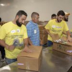 Volunteers in yellow t-shirts pack boxes labeled "Food Bank" in a busy Los Angeles Regional Food Bank warehouse. They work together on an assembly line, sealing boxes with tape and organizing supplies, ensuring everything is ready for distribution.