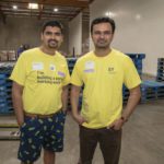 Two men wearing yellow EY shirts stand in a bustling Los Angeles Regional Food Bank warehouse. One has curly hair and the other short hair, surrounded by blue pallets with a forklift visible in the background, highlighting their commitment to alleviating hunger in the community.