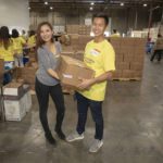 Two people smile as they hold a box at a volunteer event in the Los Angeles Regional Food Bank warehouse. Around them, other volunteers manage boxes, with shelves stocked high in the background, embodying a community spirit of giving and support.