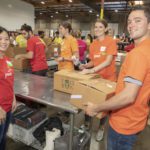 A group of volunteers wearing orange and red shirts works together in a warehouse for the Los Angeles Regional Food Bank, packing boxes labeled "Food Bank." They gather around a metal table, surrounded by shelves stocked with supplies. The atmosphere is busy and collaborative.