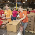 Volunteers at the Los Angeles Regional Food Bank warehouse pack boxes with food, donning colorful t-shirts, some featuring a company logo. The atmosphere buzzes with collaborative energy, amid stacks of boxes and packing materials visible in the background.