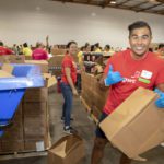 A group of volunteers at the Los Angeles Regional Food Bank is working in a warehouse, sorting cardboard boxes. One person in the foreground is smiling and giving a thumbs-up, while others in the background are actively involved in organizing and packing.
