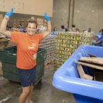 A smiling person in an orange PwC shirt and blue gloves holds up a cardboard box in the bustling Los Angeles Regional Food Bank. Surrounding them are stacks of boxes and colleagues working diligently, with a blue recycling bin in the foreground.