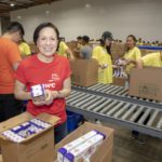 Volunteers at the Los Angeles Regional Food Bank pack food items into boxes along a conveyor belt. A woman in a red shirt holds a box of pasta, smiling at the camera, while others in yellow shirts work together tirelessly in the background.