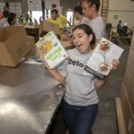 A smiling woman holds a box of corn biscuits and a brochure from the Los Angeles Regional Food Bank while volunteering in a bustling warehouse. Other volunteers are busy sorting and packing items in the background, united in their mission to support the community.