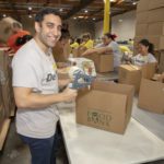 A smiling man is taping boxes at the Los Angeles Regional Food Bank while volunteering. Other volunteers are packing boxes with food items in the background, working together at tables surrounded by large cardboard boxes in a bustling warehouse setting.