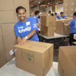 A smiling woman in a blue shirt packs boxes at the Los Angeles Regional Food Bank, surrounded by others engaged in the same effort. Stacks of cardboard boxes fill the background, creating a busy and productive atmosphere.