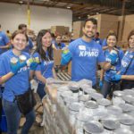 In a bustling warehouse, volunteers in blue shirts and gloves from the Los Angeles Regional Food Bank hold canned goods, beaming at the camera. Behind them, stacks of canned food and cardboard boxes showcase their dedication to fighting hunger in the community.