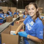 A group of volunteers in blue shirts packs boxes at the Los Angeles Regional Food Bank. A woman in the foreground smiles while placing items in a box. The background shows others working along a conveyor belt in the large warehouse.