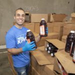 A man in a blue shirt and gloves smiles while holding a large bottle of juice, standing by several open cardboard boxes filled with similar bottles at the Los Angeles Regional Food Bank warehouse.