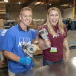 Two people smiling and holding tape guns in a bustling warehouse, likely at the Los Angeles Regional Food Bank. They stand before stacked cardboard boxes, with one wearing a blue KPMG shirt and the other in a maroon PWC shirt with a name tag, embodying teamwork and community spirit.