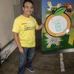 A person in a yellow T-shirt stands smiling next to a sign shaped like an orange, displaying "4,972 total boxes" at the Los Angeles Regional Food Bank. Behind them, a metal shelving unit is visible in the warehouse setting.