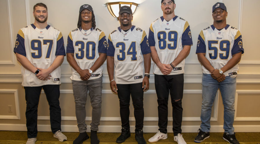 Five men in matching sports jerseys pose indoors, standing side by side in front of a beige wall and a sconce. They are casually dressed, wearing jeans and sneakers. Representing the Los Angeles Regional Food Bank, their jerseys feature a blue and gold color scheme with numbers on the front.