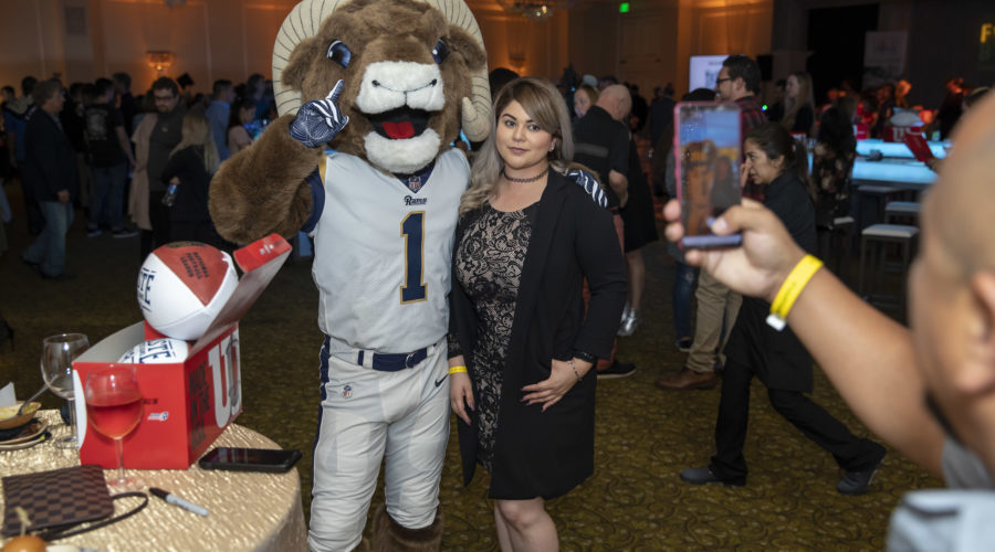 A person poses with a costumed mascot resembling a ram at an indoor event supporting the Los Angeles Regional Food Bank. The mascot sports a football uniform, while another individual in the foreground snaps a photo on their smartphone. Attendees and various event elements are visible in the background.