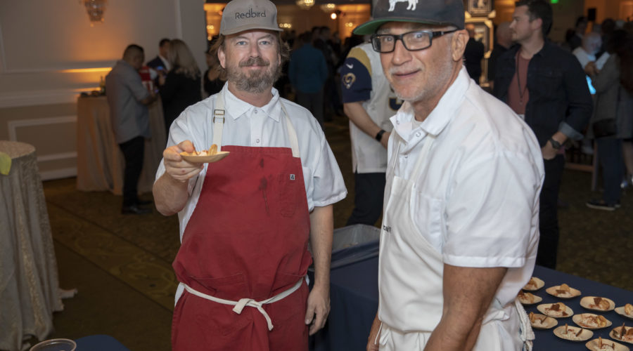 Two chefs wearing aprons and hats serve food at a Los Angeles Regional Food Bank event. One chef holds a plate with appetizers, while the other stands beside him behind a table with more dishes. People are gathered in the background, enjoying the culinary offerings.