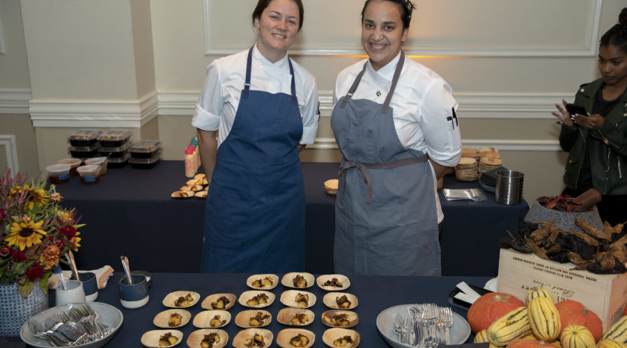 Two chefs wearing aprons stand smiling behind a table laden with gourmet food, small pumpkins, and flowers. They're collaborating with the Los Angeles Regional Food Bank. In the background, a person looks at a phone.