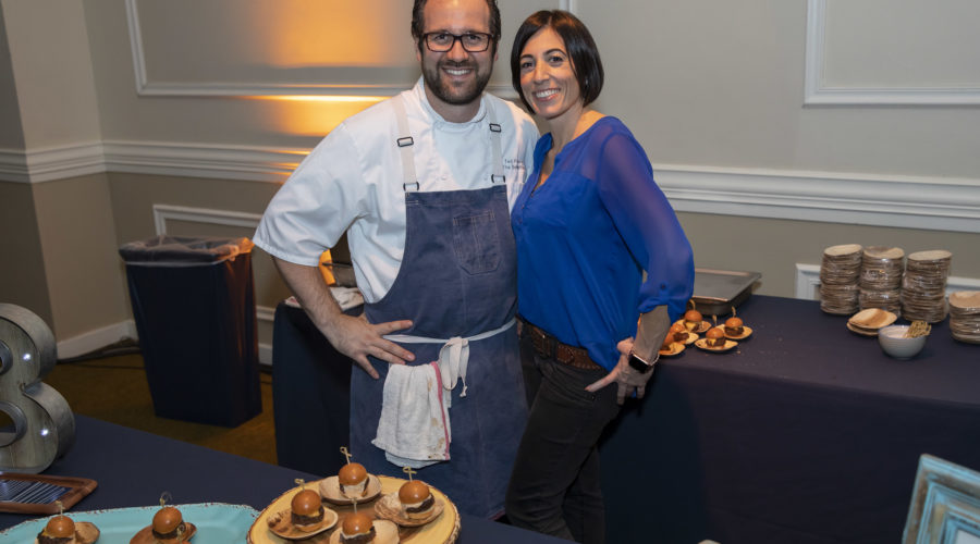 A man in a chef's uniform and a woman in a blue blouse are smiling and posing together at an indoor event for the Los Angeles Regional Food Bank, standing proudly in front of a table filled with small burgers on wooden platters.
