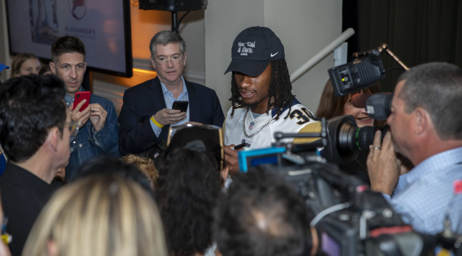 A man in a cap signs autographs for a crowd of fans and media personnel at what seems to be an indoor event supporting the Los Angeles Regional Food Bank. People surrounding him take photos with their phones, while a cameraman captures the bustling scene.