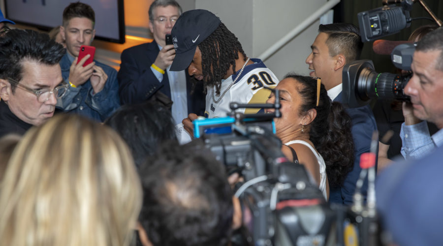 In a crowded room brimming with excited fans and media, a sports player wearing a cap and jersey with "30" signs autographs. Camerapersons and onlookers capture the moment, as he shares stories about his recent visit to the Los Angeles Regional Food Bank.