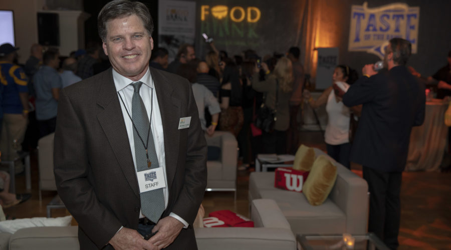 A man in a suit stands smiling at an indoor event, wearing a badge labeled "staff." Behind him, groups of people socialize as colorful banners for the Los Angeles Regional Food Bank and "Taste of the NFL" adorn the walls. The setting is casual yet organized.