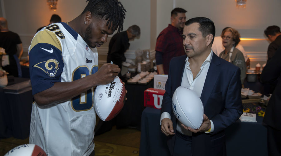 A football player in a Rams jersey autographs a football for a man in a suit at an indoor event supporting the Los Angeles Regional Food Bank. Other attendees and event displays are visible in the background, highlighting the community's effort.