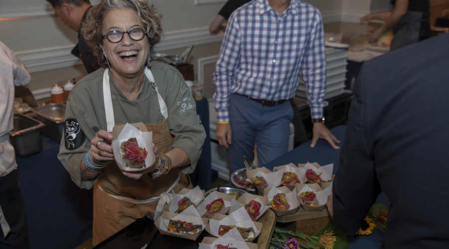 A woman wearing glasses and an apron, representing the Los Angeles Regional Food Bank, smiles as she holds up food wrapped in paper. Trays filled with similar items are in front of her, while others mingle in the background at this bustling indoor event.