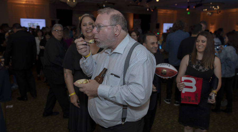 At a crowded party, a man in a gray shirt and suspenders enjoys food reminiscent of Los Angeles Regional Food Bank offerings. Around him, guests hold items like a football and a red folder, while the dimly lit room glows softly under the chandelier above.