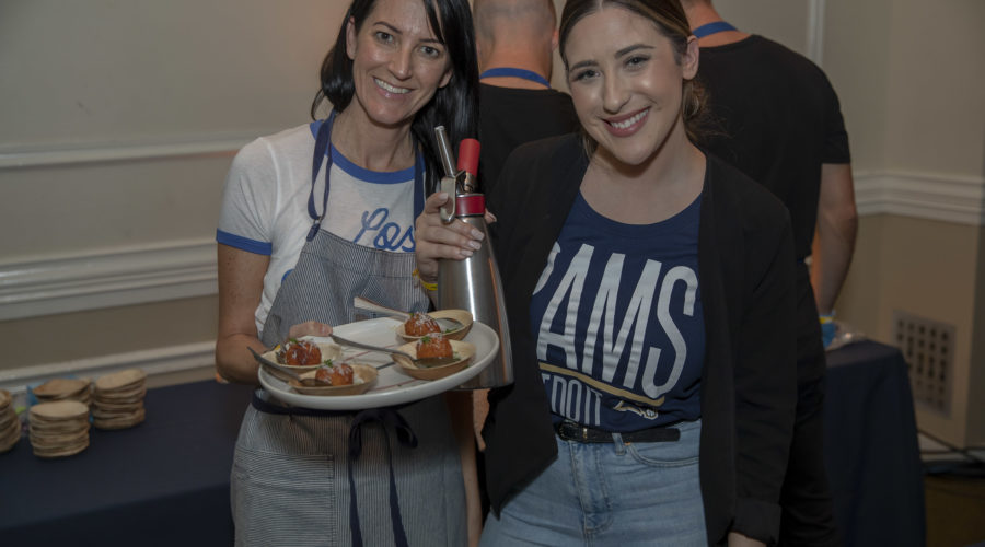 At a vibrant food event, two women beam with joy. One, donning an apron, holds a plate of appetizers and a whipped cream dispenser. Her friend stands beside her in a black jacket and T-shirt. The lively scene could easily be part of a Los Angeles Regional Food Bank gathering with others blurred in the background.