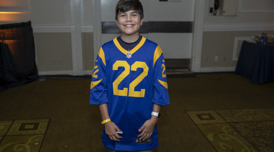 A smiling person stands indoors wearing a blue and yellow sports jersey with the number 22, supporting the Los Angeles Regional Food Bank. They are in a room with beige walls, patterned carpet, and a dark blue draped table in the background.