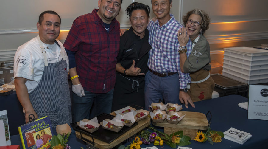 A group of five people stands behind a table adorned with decorative flowers and trays of food samples. They're smiling, radiating a relaxed and friendly atmosphere at an event supporting the Los Angeles Regional Food Bank.