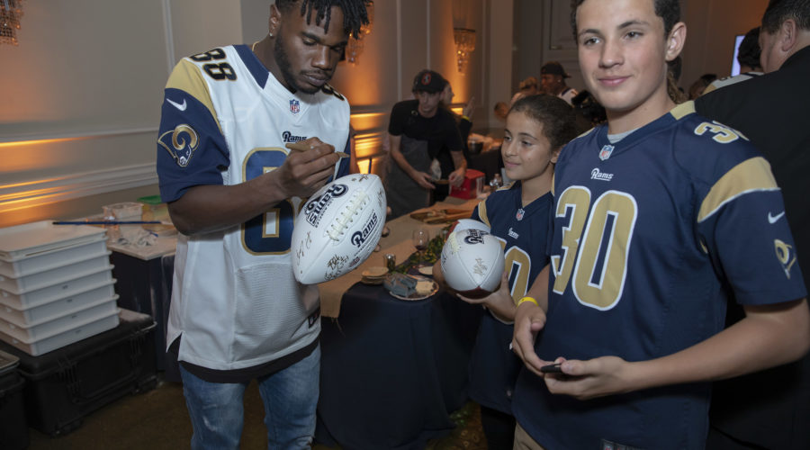 A football player in a jersey signs a football for two young fans wearing team jerseys at the Los Angeles Regional Food Bank event. They are indoors, with other people and tables visible in the background.