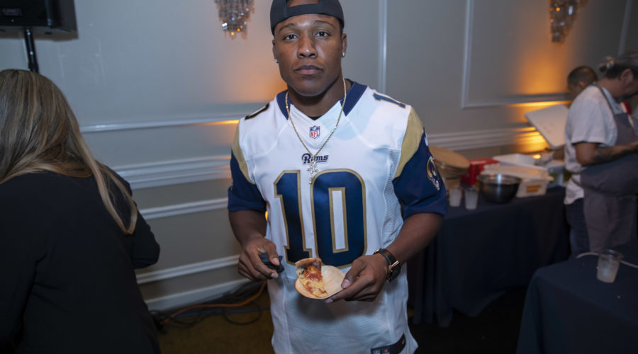 A person in a white and blue sports jersey stands in a dimly lit room adorned with chandeliers, holding a slice of pizza and a phone. The lively gathering, which supports the Los Angeles Regional Food Bank, buzzes with energy as others mingle in the background.
