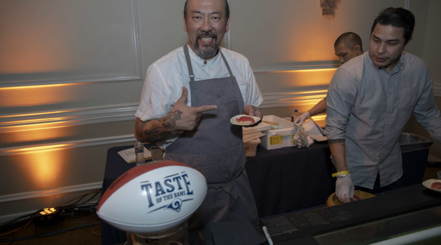 A chef wearing an apron smiles and points at a plate of food in his hand, showcasing a dish prepared for the Los Angeles Regional Food Bank. A football labeled "Taste of the NFL" is displayed in the foreground. Another person stands nearby at a table with cooking ingredients. The room is warmly lit.