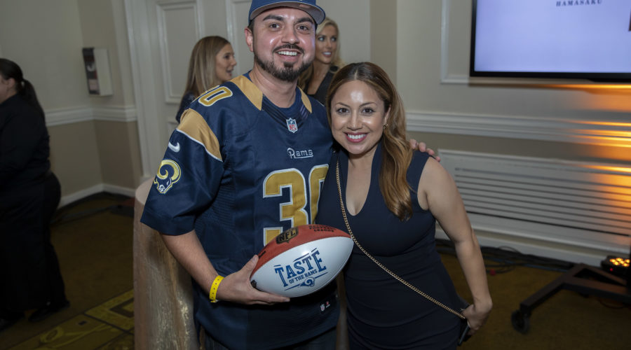 A man in a Los Angeles Rams jersey holds a football while posing with a woman in a black dress. They're indoors at an event supporting the Los Angeles Regional Food Bank, with a TV screen displaying the words "UMI HANASUKE" in the background.