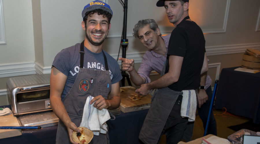 Three men in aprons and caps are smiling and preparing food in a kitchen setting. One is holding a tortilla while proudly wearing a "Los Angeles" T-shirt, embodying the casual and cheerful spirit of the Los Angeles Regional Food Bank.
