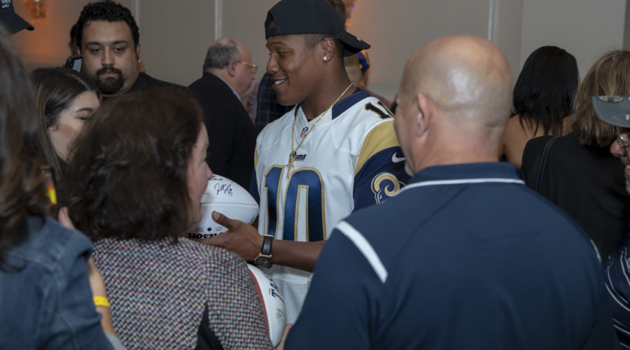 In a bustling indoor setting with chandeliers overhead, a man in a blue sports jersey and cap smiles warmly as he signs a football helmet. He's engaging with the crowd at an event supporting the Los Angeles Regional Food Bank.