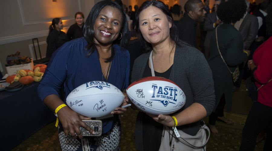 Two women smile, holding autographed footballs at an indoor event benefiting the Los Angeles Regional Food Bank. The setting is lively, with delicious food and guests mingling in the background. The footballs proudly display "Taste of the Rams.