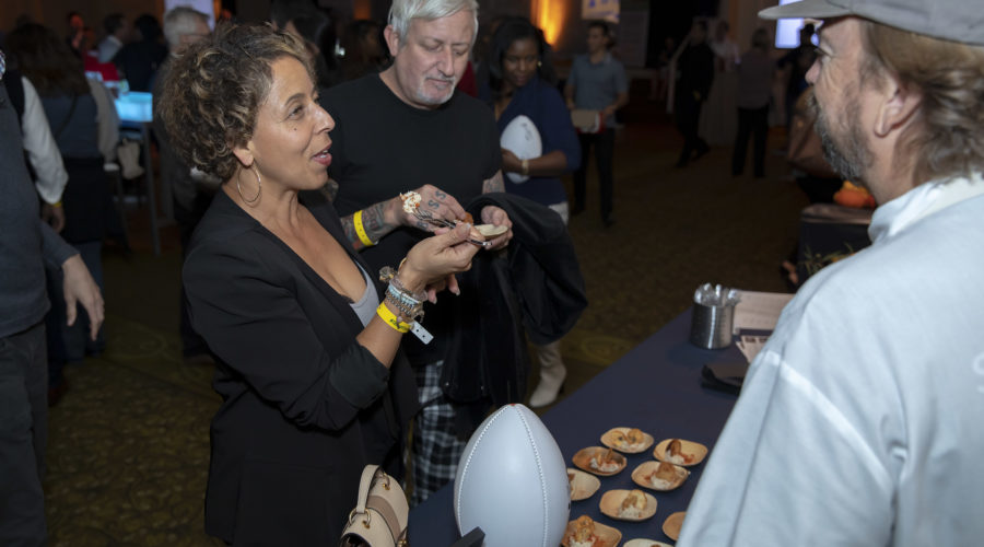 A woman with short curly hair chats with a man in a cap at a booth, surrounded by small plates of food and a football-shaped object. Nearby, another man with gray hair stands watchfully. The lively setting hints at an indoor event like the Los Angeles Regional Food Bank gathering.