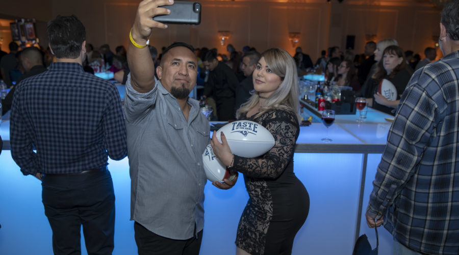 A man and a woman pose for a selfie at an event supporting the Los Angeles Regional Food Bank. The woman holds a football with the word "TASTE" on it, as people mingle in the background near a bar illuminated by blue lighting.