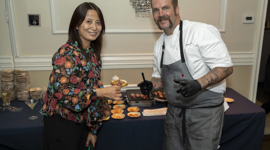 A woman in a floral blouse holds a plate of food, smiling at the camera next to a chef in a white shirt and apron. The chef, supporting the Los Angeles Regional Food Bank, is serving dishes on a table with a navy tablecloth featuring various food items. They stand in an elegantly decorated room.