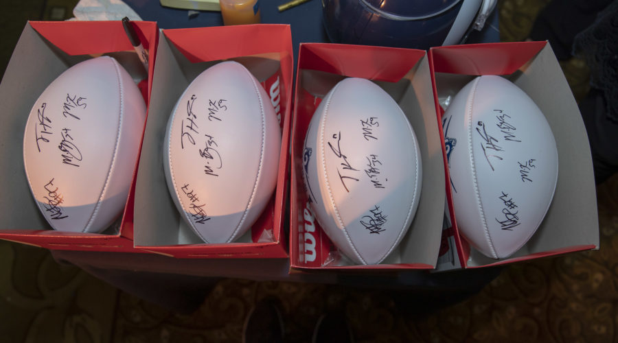 Four autographed footballs are displayed in opened boxes on a surface, each partially covered by its box lid. A helmet and a bottle from the Los Angeles Regional Food Bank are visible in the background, adding a touch of community spirit to the scene.