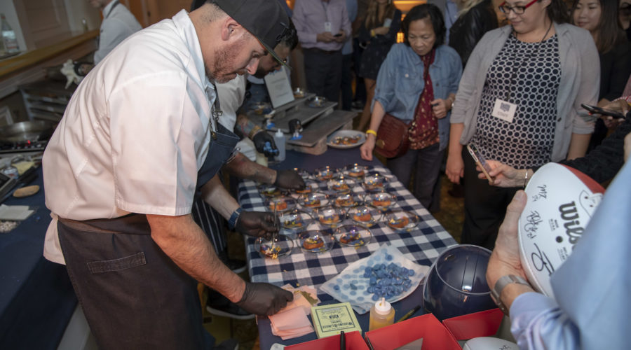 A chef in a white shirt and apron is preparing food at a table covered with dishes. A group of people, some holding phones, watches him intently. Nearby, autographed footballs in red boxes add to the excitement of this Los Angeles Regional Food Bank event.